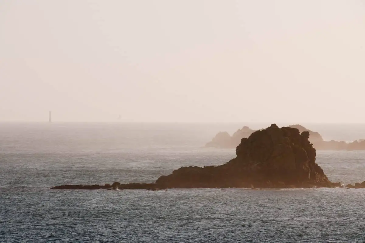 Rocky formations emerging from the calm sea under a hazy, light sky. A small lighthouse is visible in the distance, creating a serene and atmospheric seascape.
