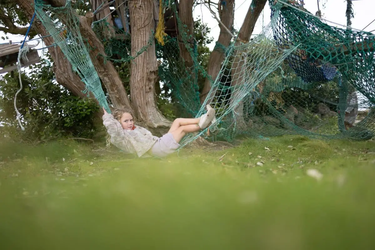 Child relaxing in a hammock made of blue netting, tied between trees. The area is grassy with scattered leaves. The child is wearing a sweater, shorts, and sneakers, surrounded by a wooded environment.