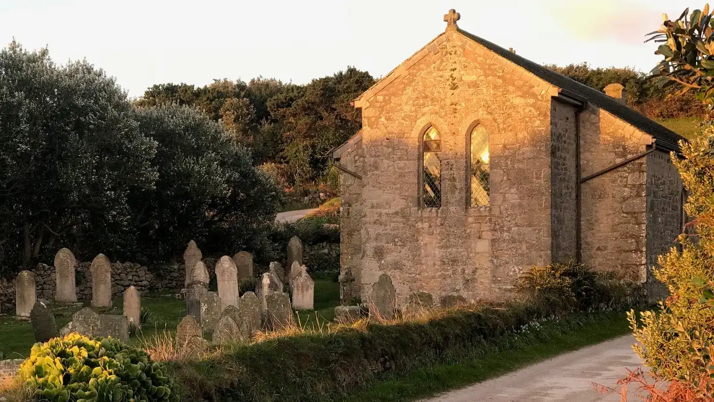 A small stone church with arched windows stands beside a cemetery with old gravestones, surrounded by greenery and lit by warm, golden sunlight.