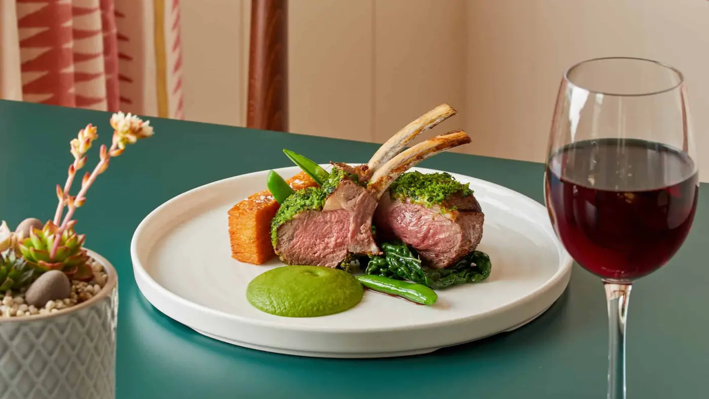 A plated dish of herb-crusted lamb chops with vegetables and a green puree sits on a teal table. To the side, a glass of red wine and a small potted plant with succulents. The background features a patterned curtain.
