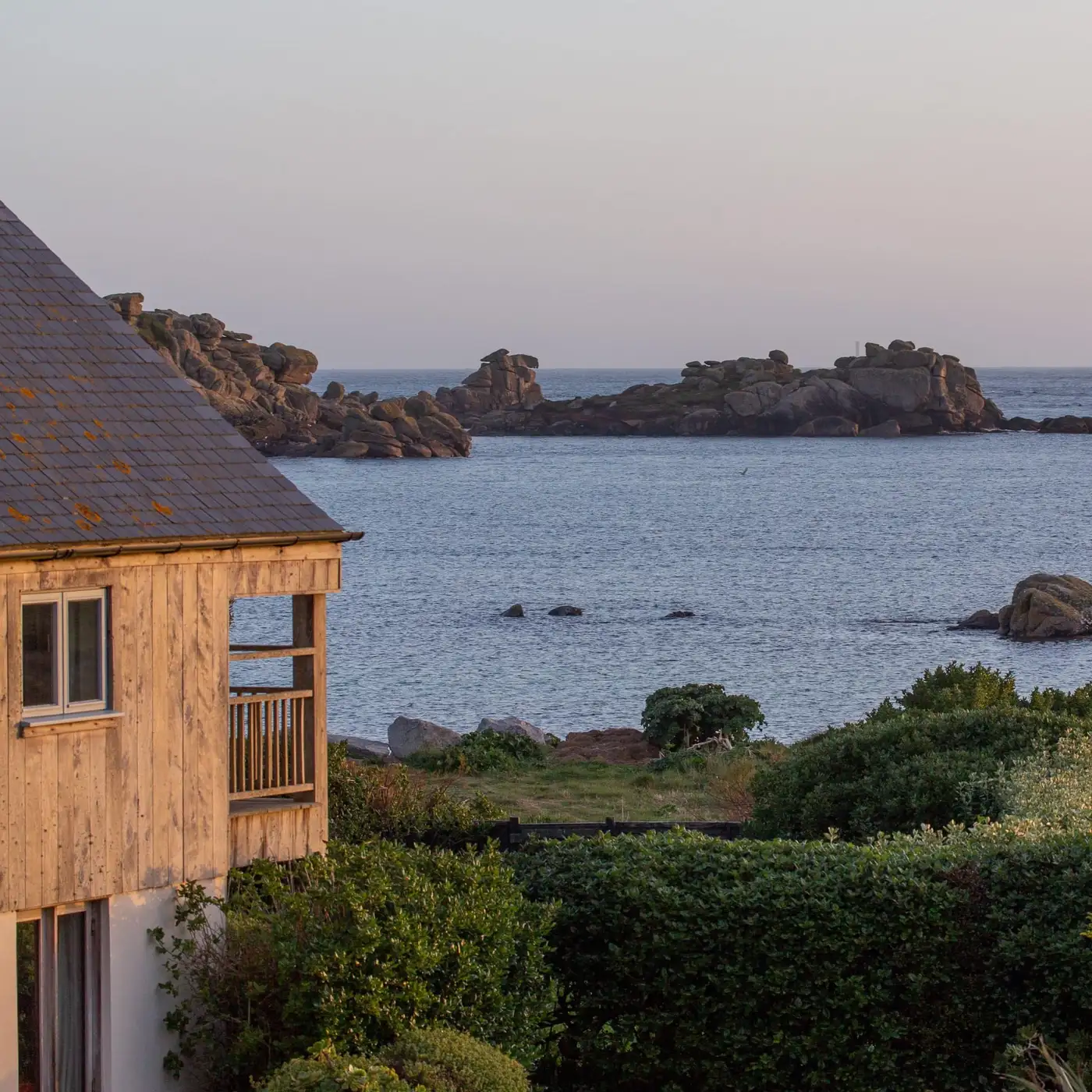The wood-clad building of Juno bedroom with a balcony is surrounded by greenery, overlooking a calm sea with rocky outcrops under a pastel-colored sky at sunset.