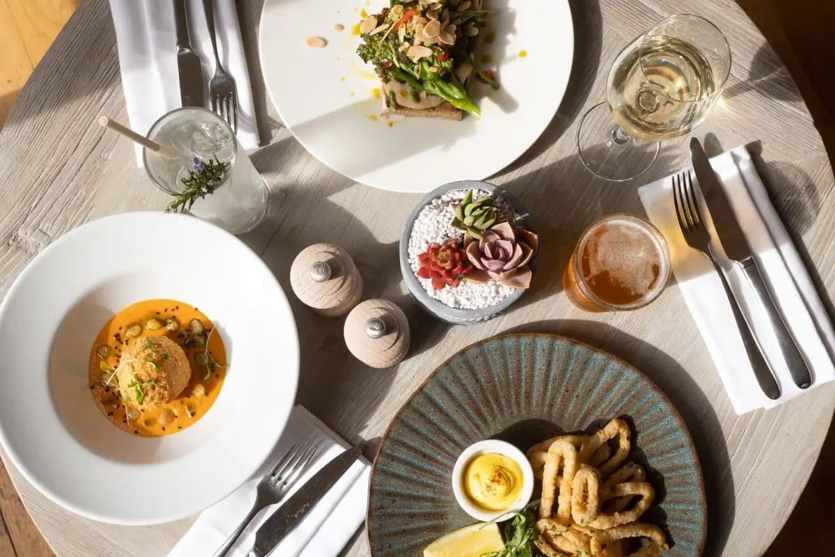 A round table with four dishes: a seafood dish in tomato sauce, asparagus with nuts on a white plate, calamari with a boiled egg on a blue plate, and a small bowl of decorative plants. Drinks include white wine, beer, and a cocktail.