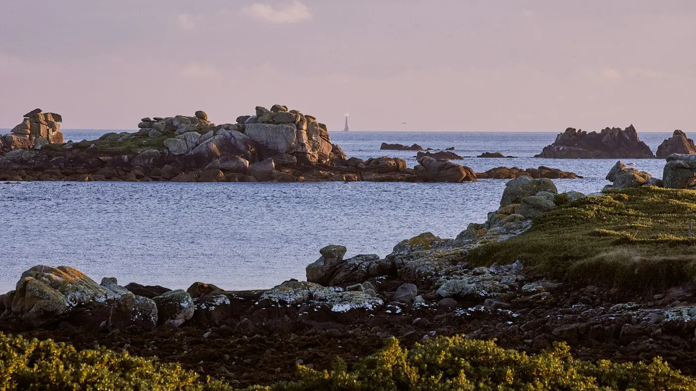 Rocky coasts and grassy areas frame a calm sea in the foreground. In the distance, a lighthouse is visible on the horizon under a soft, cloudy sky.