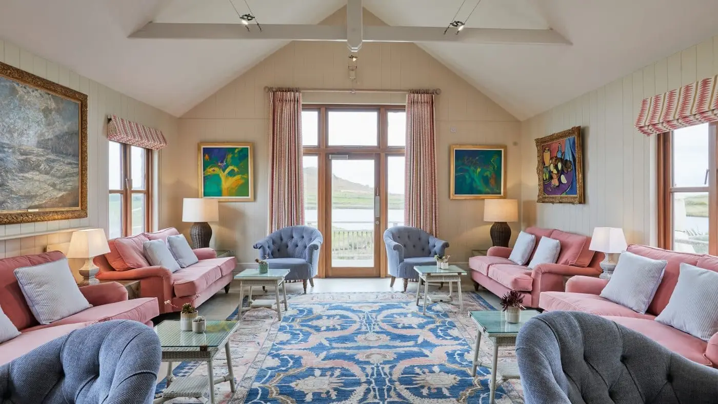 A spacious lounge features pink sofas, blue armchairs, and a large patterned rug. Paintings decorate the cream walls, and a tall window offers a scenic view outside. Decorative cushions and table lamps add to the cosy ambiance.