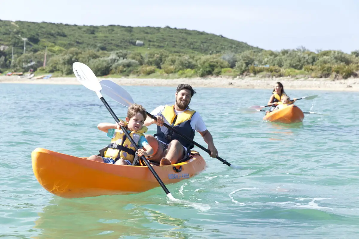 A man and a child paddle together in an orange kayak on clear turquoise water, followed by a woman in another kayak. They all wear life jackets, and a green, hilly shoreline is visible in the background under a clear sky.