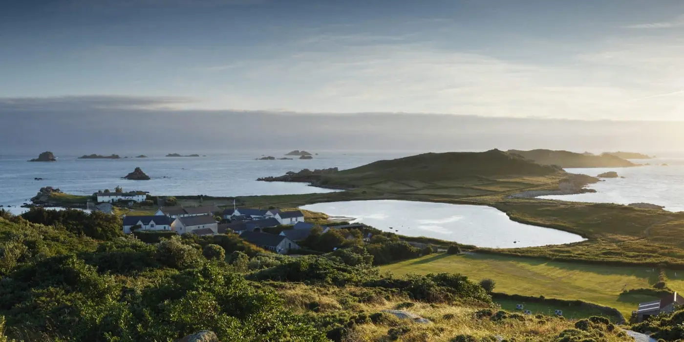 A scenic view of a coastal landscape with a collection of buildings by a lagoon. There are green fields, rolling hills, and scattered islands in the background under a partly cloudy sky.