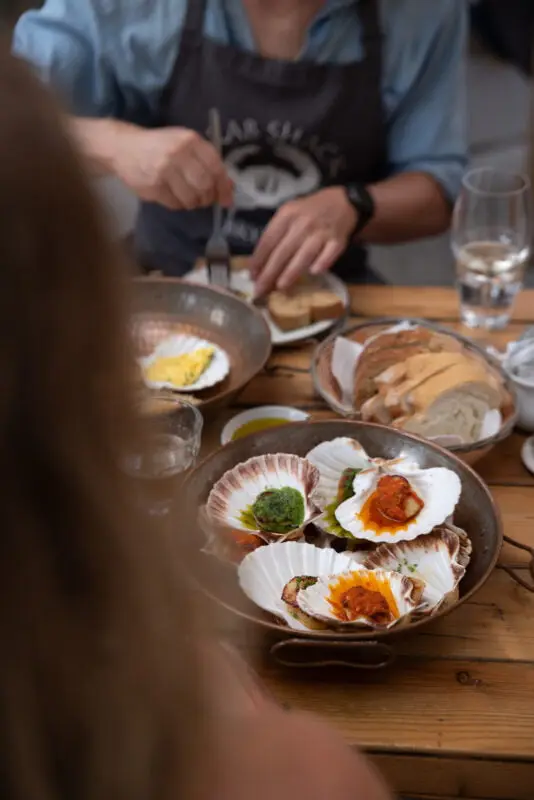 A person is seated at a wooden table with several plates of food, including scallops on shells with various toppings, sliced bread, and dipping sauces. The focus is on the scallops, with the person partially visible in the background.