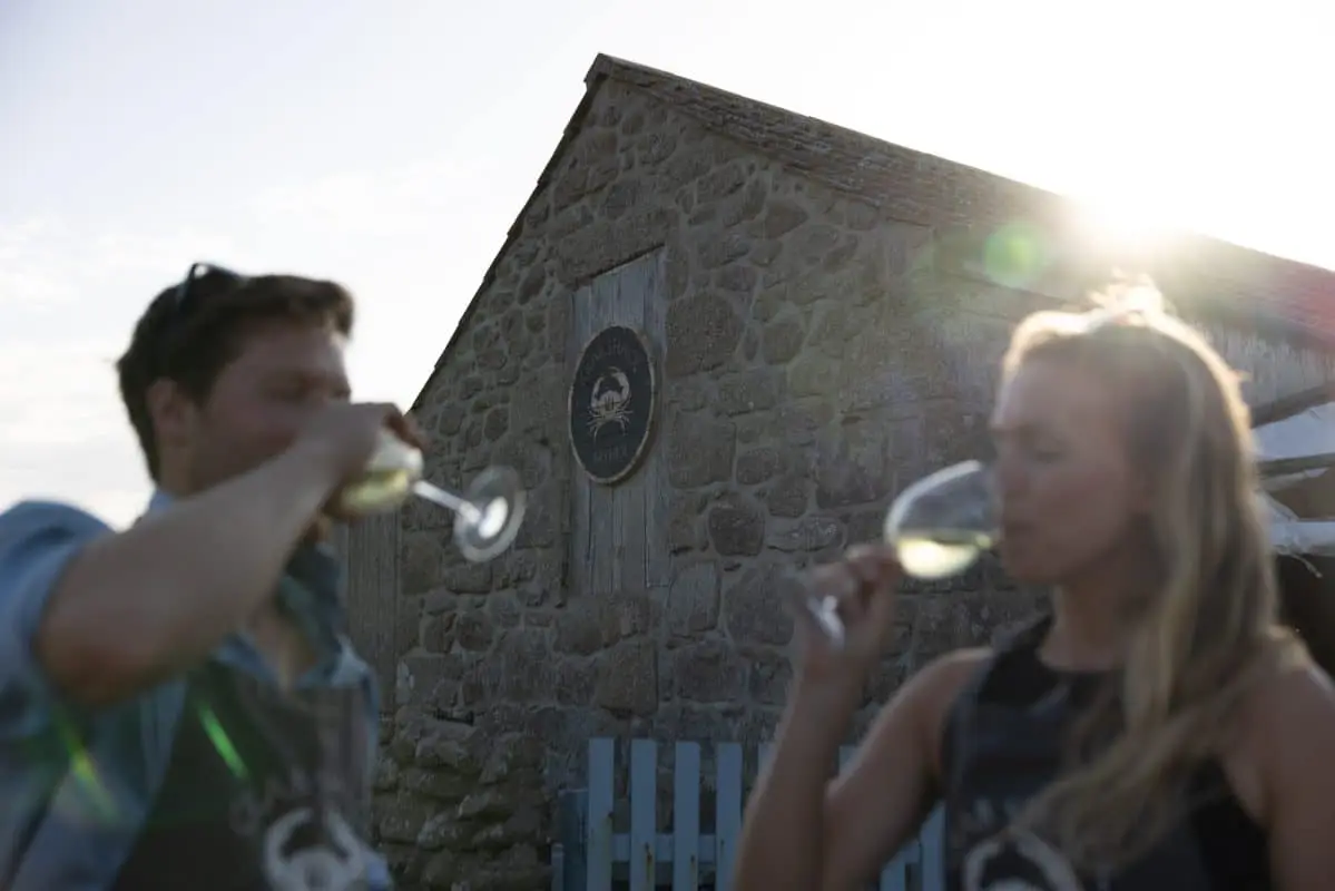 Two people sip white wine in front of a rustic stone building with a crab emblem on the wall. Bright sunlight creates a warm glow around them.