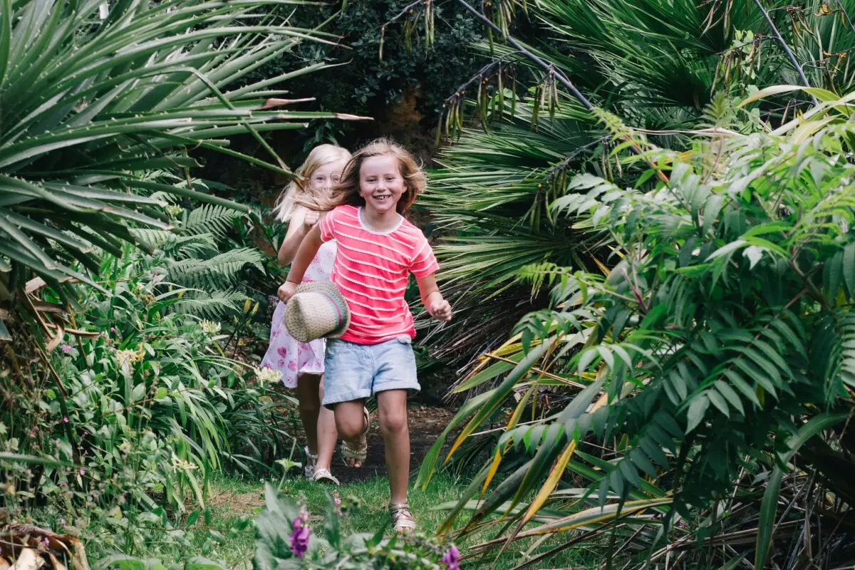 Two children joyfully run through a lush, green garden path. The girl in front holds a straw hat and wears a red striped shirt and blue shorts. The girl behind wears a floral dress. Tall, verdant plants surround them.