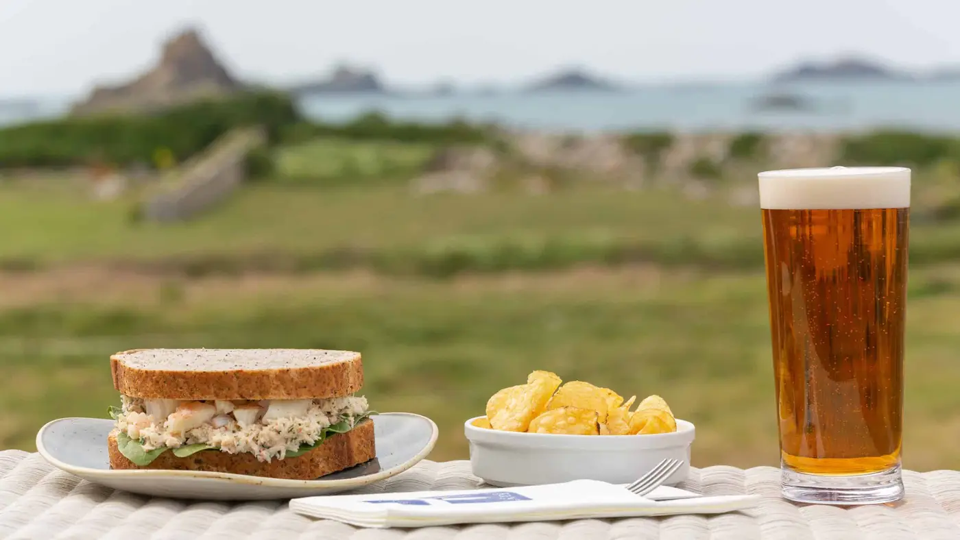 A close-up of a meal on a patio table with a scenic background. The meal includes a sandwich on whole-grain bread, a bowl of potato chips, and a glass of beer. The backdrop features grassy fields, rocky formations, and a calm sea.