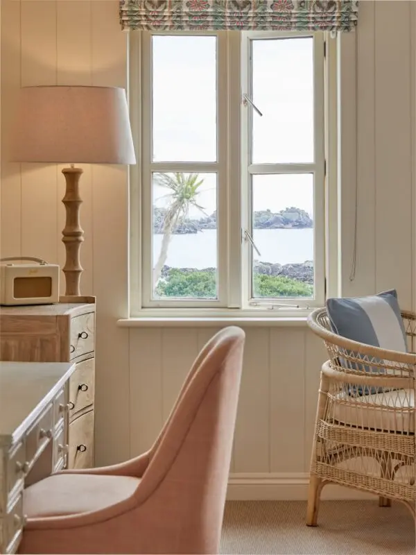 A cosy room featuring a desk with a lamp, a pink upholstered chair, and a wicker chair with a cushion. The window offers a view of the sea, rocks, and a palm tree outside. Natural light illuminates the interior.