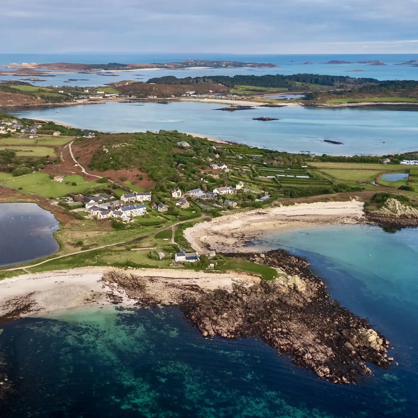 Aerial view of Bryher with Popplestones and Great Par beaches visible with a blue and turquoise sea surrounding the land, dotted with small islands under a partly cloudy blue sky.