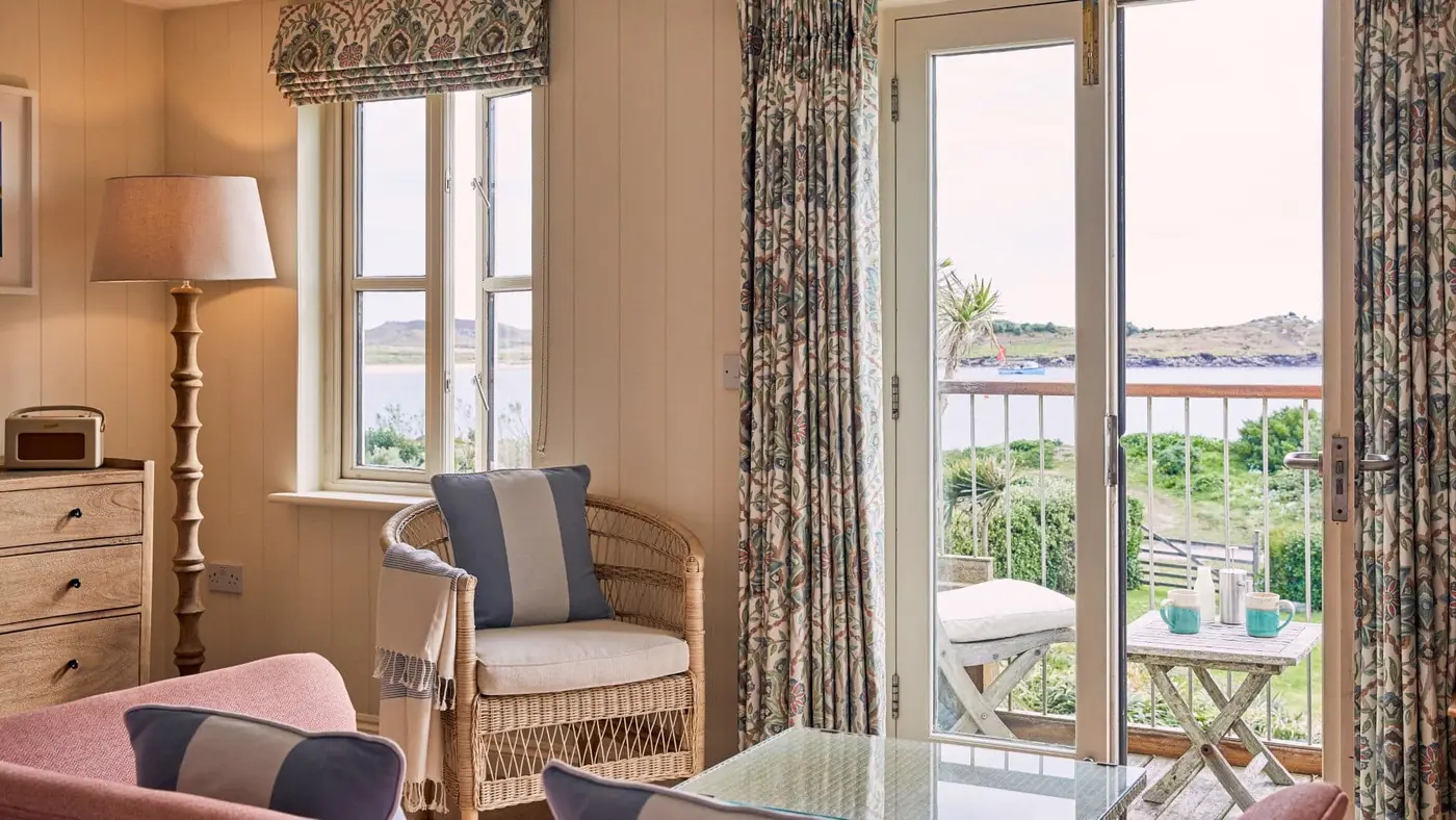 Cosy living room with wicker chair and glass table, overlooking a balcony with a scenic view of the ocean and greenery. Tall lamp and patterned curtains frame the windows, creating a warm, inviting atmosphere.