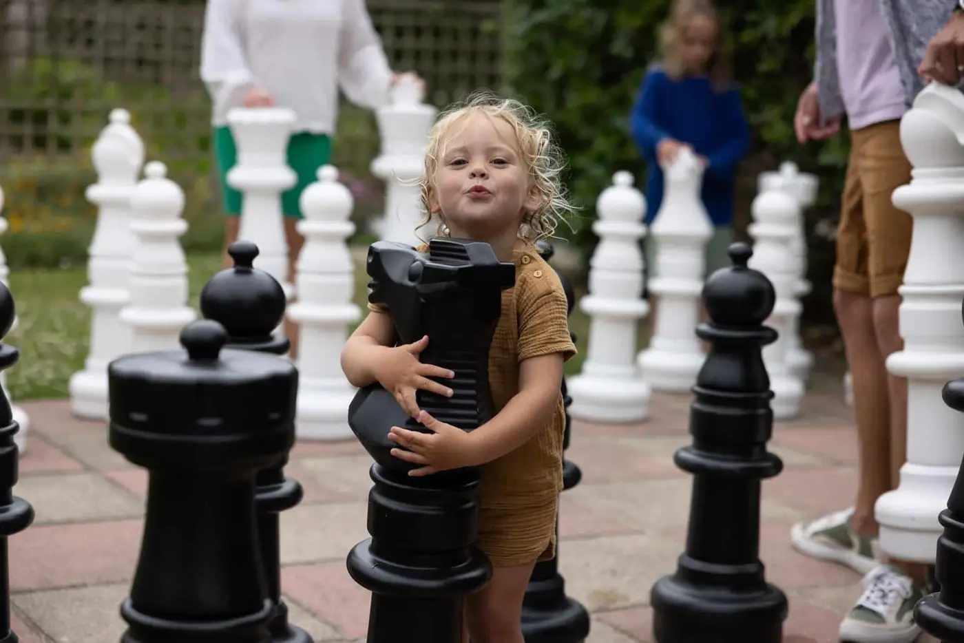 A young child with blonde curls hugs a large black knight chess piece on an oversized outdoor chessboard. People in the background are interacting with other giant chess pieces, both black and white. The setting is casual and playful.