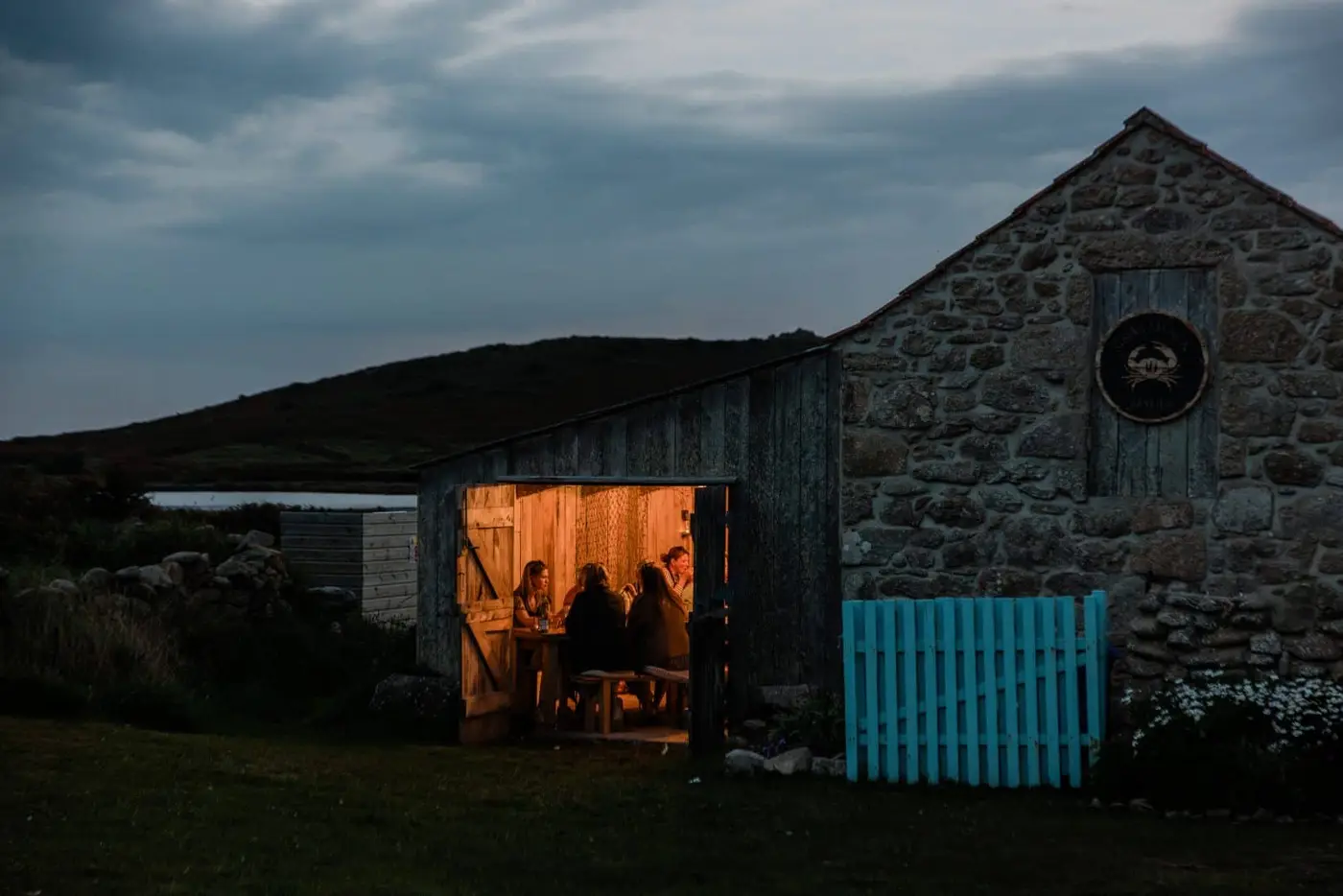 A stone building glows warmly from inside, contrasting with the darkening evening outside. Visible through the open door, several people are gathered around a table. A blue wooden gate and a hilly landscape are in the background.