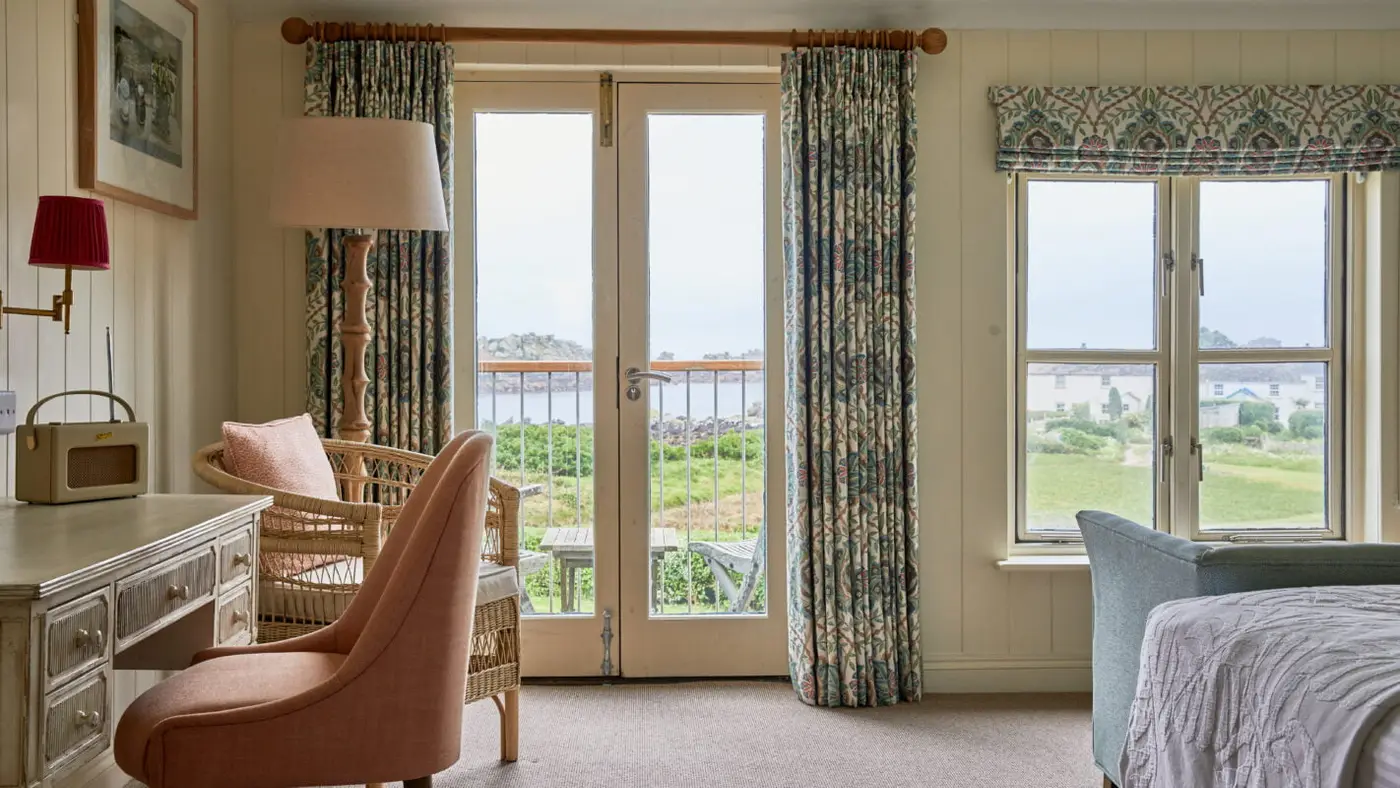 Cosy room with a desk, pink chair, wicker armchair, and lamp. French doors and large window with patterned curtains frame a scenic view of greenery and the ocean. Bed with partial green armchair visible. Neutral and pastel tones dominate.