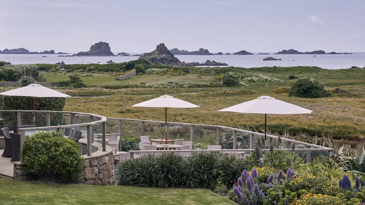A scenic coastal view features a garden with white umbrellas and seating behind a glass fence. In the background, rocky formations rise from the sea under a cloudy sky, surrounded by lush greenery and colourful flowers.