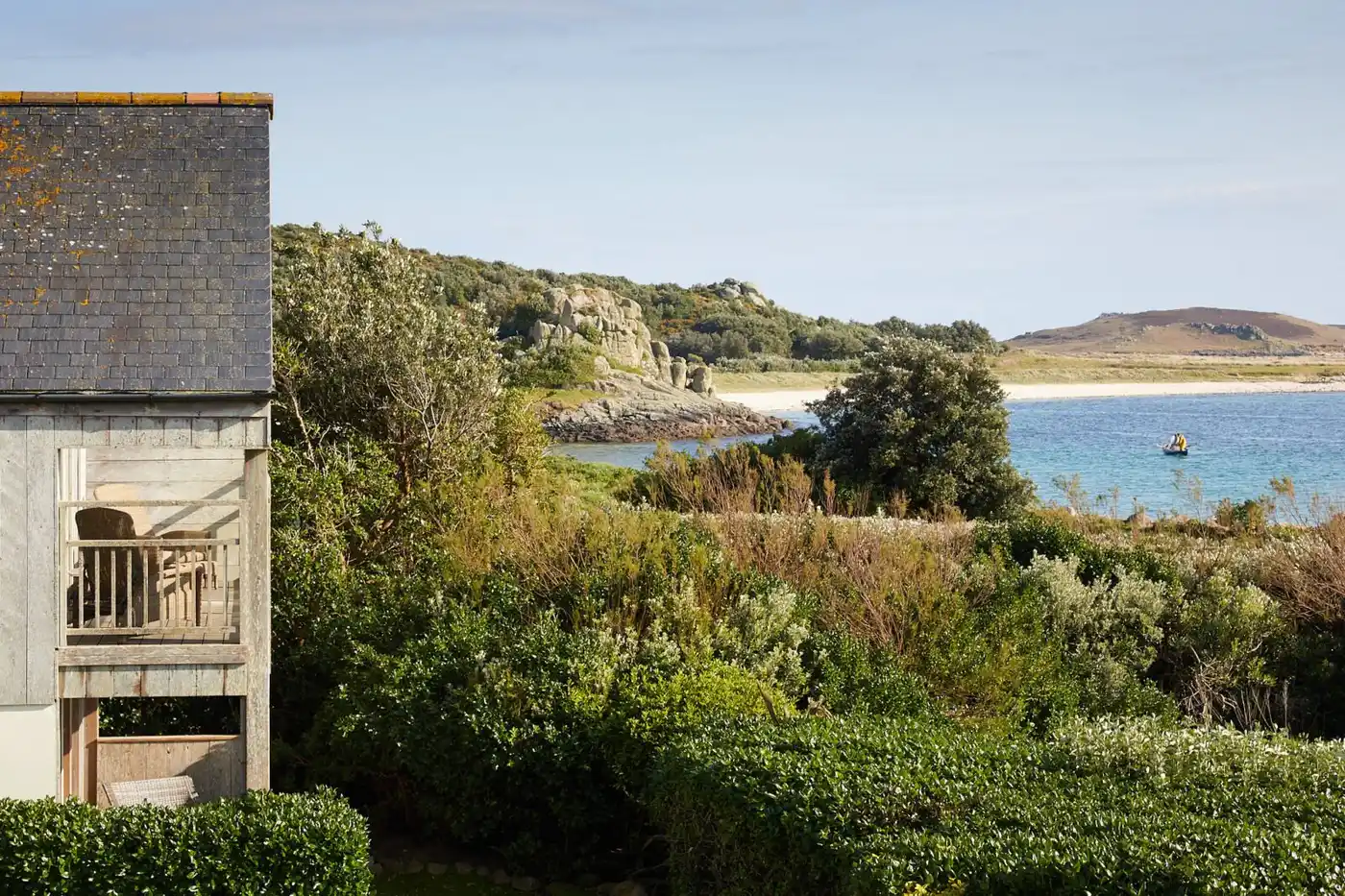 A scenic view of a coastal landscape with the wooden-clad Juno bedroom building visible on the left. It features a wooden balcony overlooking lush greenery and a sandy beach leading to the blue ocean. A small boat is on the water, and hills are seen in the background.