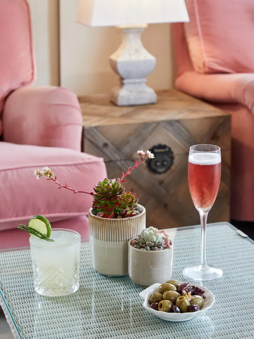 A cosy lounge scene featuring a glass of pink champagne and a cocktail garnished with lime on a glass table. There are two succulents in pots and a bowl of olives with sundried tomatoes. In the background, there are a pink armchair and a wooden lamp.