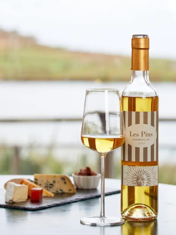 A bottle of Les Pins Monbazillac 2021 wine is next to a filled wine glass on a table. In the background, theres a platter with assorted cheeses and a scenic view of a river and landscape in soft focus.
