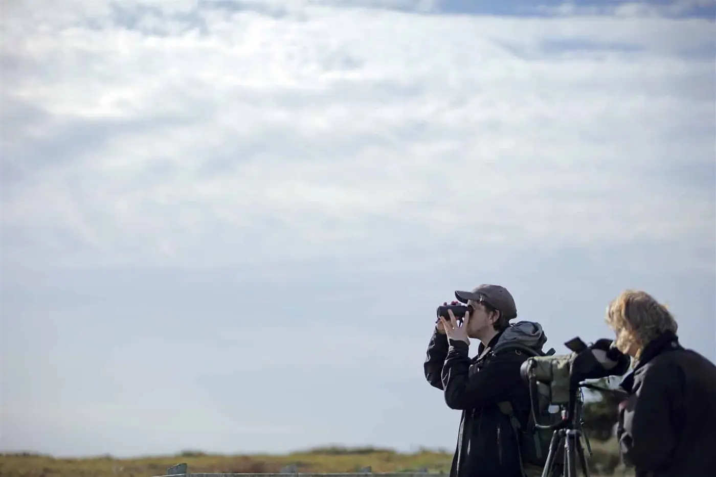 Two people outdoors, one looking through binoculars and the other adjusting a camera on a tripod. They are dressed warmly, standing under a partly cloudy sky, with grassy fields in the background.