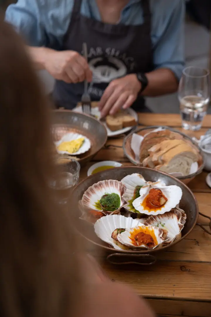 A person is seated at a wooden table with several plates of food, including scallops on shells with various toppings, sliced bread, and dipping sauces. The focus is on the scallops, with the person partially visible in the background.