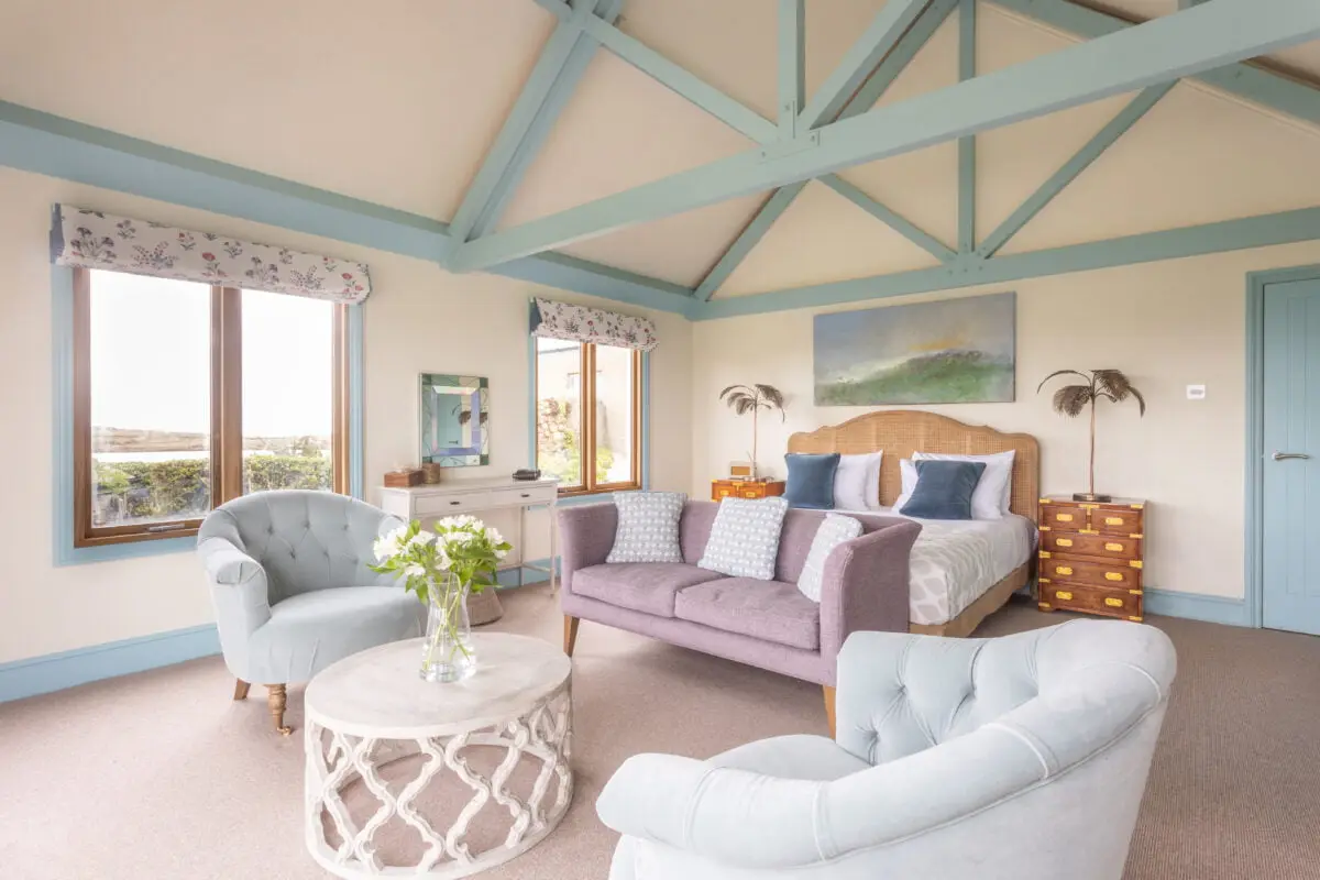 A cosy bedroom with a vaulted ceiling featuring pastel blue beams. There's a large bed with pillows, a sofa, and two cushioned chairs. A round coffee table with flowers is in the foreground. Light streams in through two windows with floral curtains.