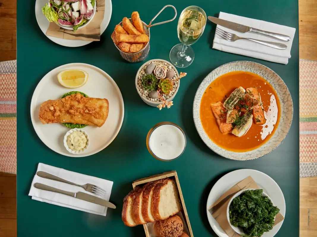 A green table set for a meal, featuring battered fish with a lemon wedge, chips in a basket, crusty bread, a salmon dish in sauce, sides of salad and kale, a pint of beer, and a glass of white wine.