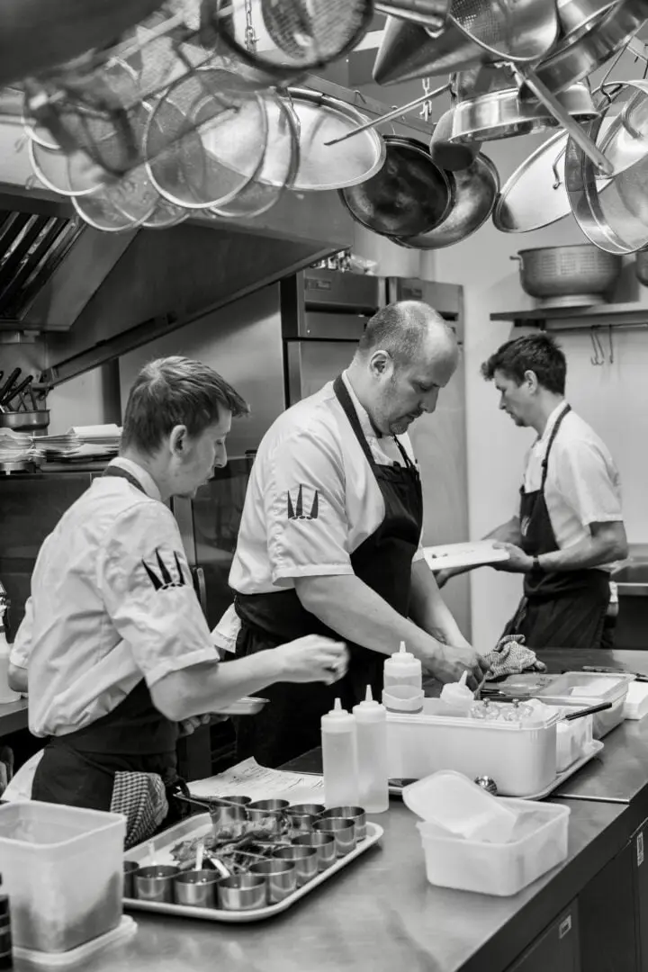 Three chefs work together in a commercial kitchen. One is preparing food on a counter, while the others assist nearby. The kitchen is equipped with various pots, utensils, and containers. The scene is in black and white.