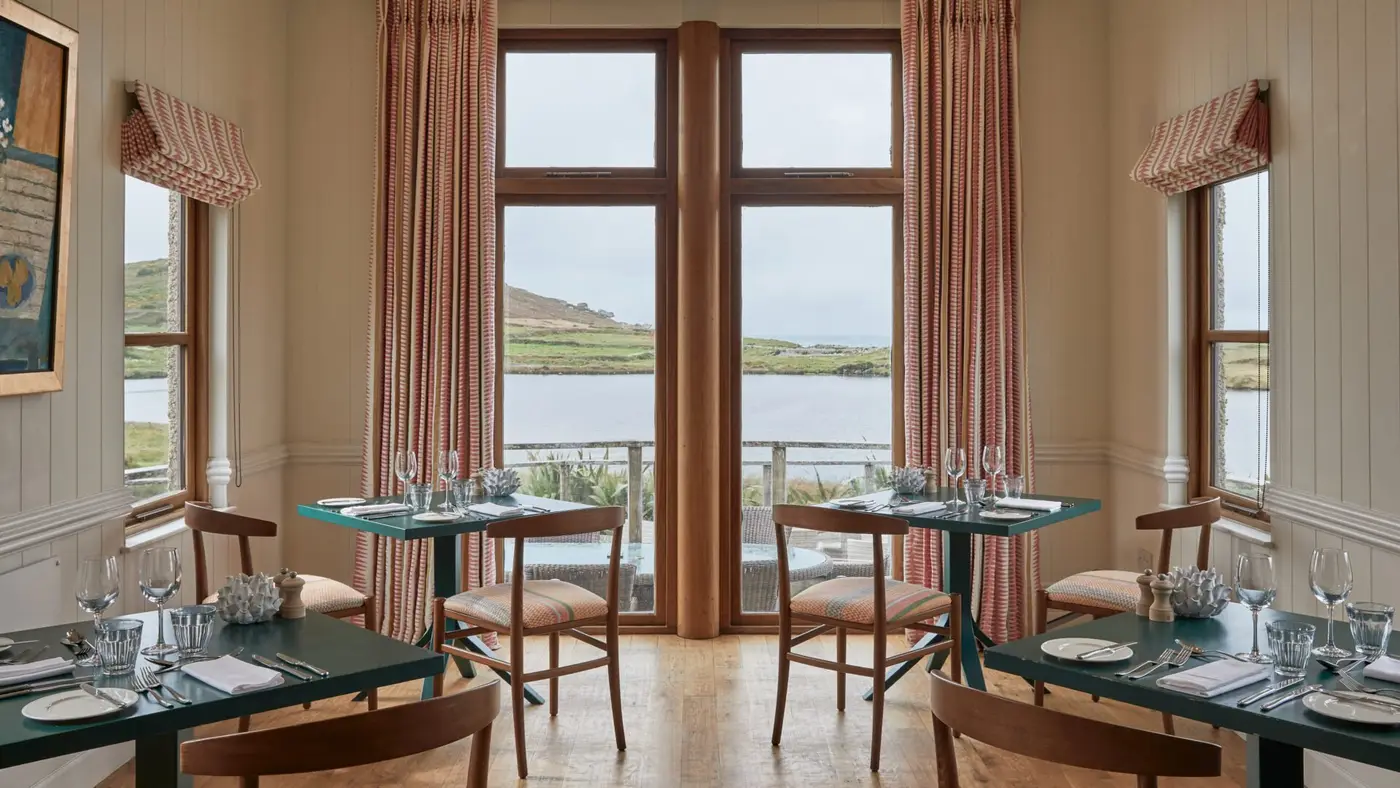 A cosy dining room with wooden tables and chairs, set with glassware and cutlery, overlooks a serene ocean view. Tall windows with striped curtains frame the scenic landscape of green hills and water. Soft lighting enhances the warm atmosphere.