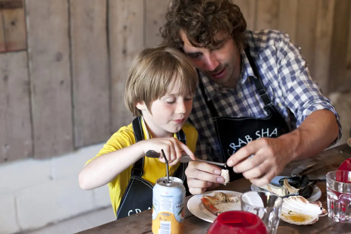 A man and a child wearing aprons are sitting at a table with seafood. The child is holding a piece of shell, while the man helps. A drink can and seafood shells are on the table. The background is wooden.