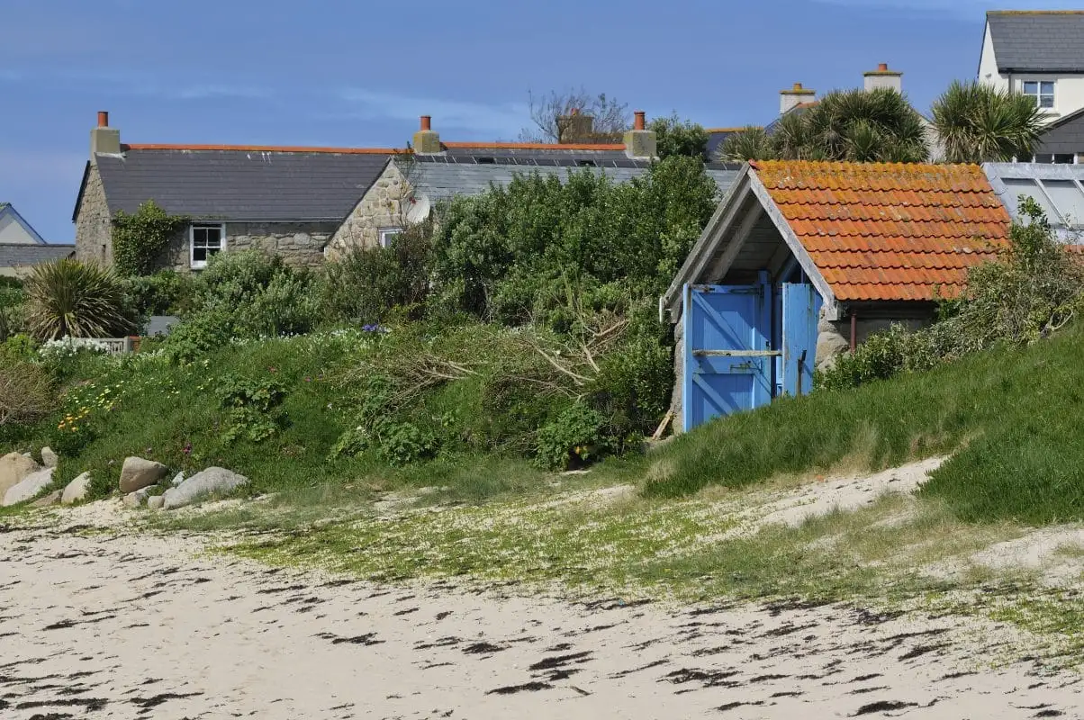 A sandy beach with some seaweed leads to a grassy area with a small, rustic former gig shed with a red tile roof and blue door. In the background are stone houses with chimneys, surrounded by lush greenery and a blue sky overhead.