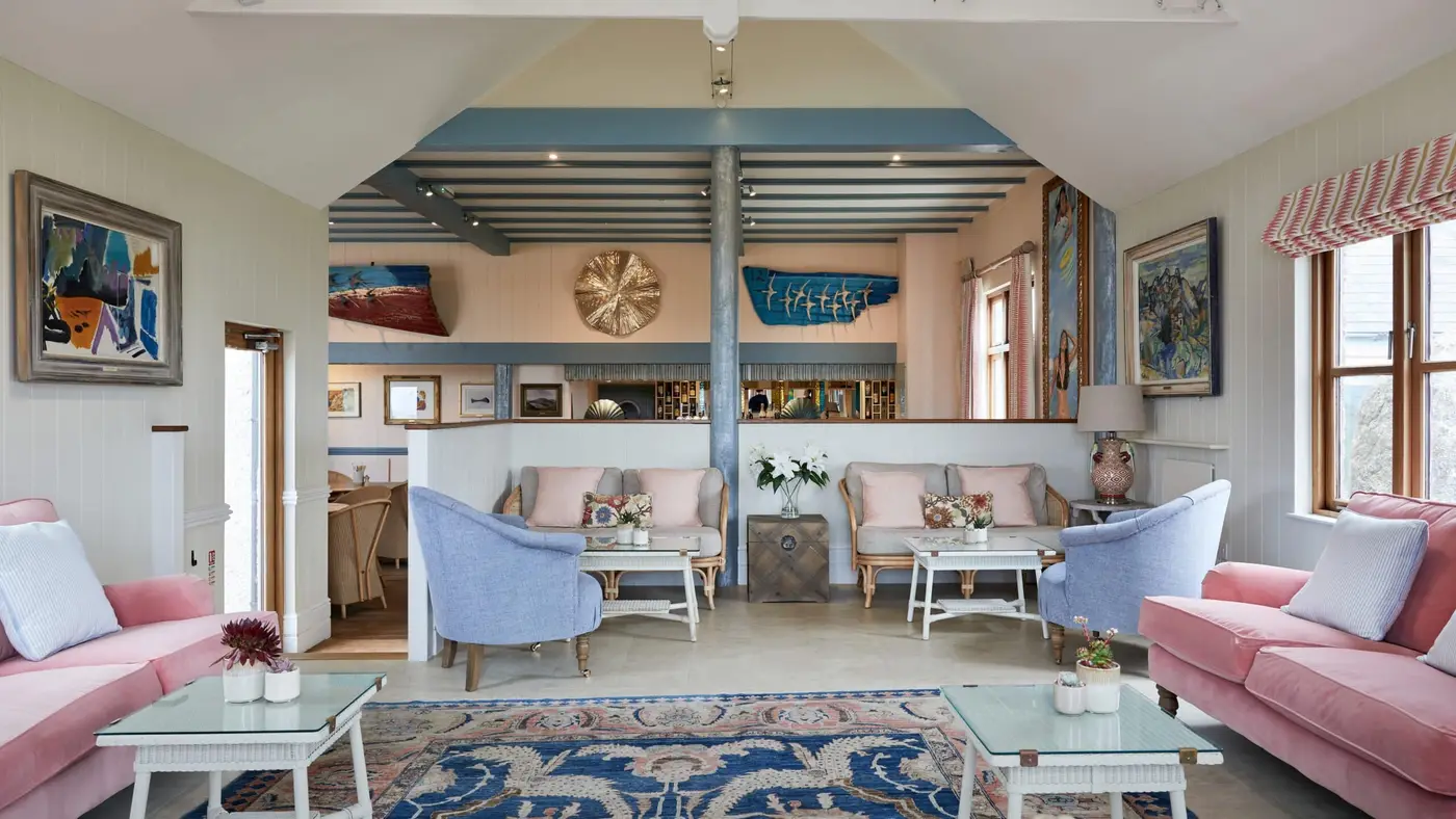 The Lookout Lounge at Hell Bay - a cosy space featuring pastel-coloured sofas and armchairs, a patterned rug, and various paintings on the walls, with a Bar visible in the background. Large windows let in natural light, and there are flowers on the tables.