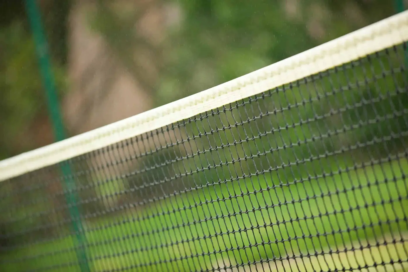 Close-up view of a tennis net with a blurred background of green grass and trees. The net, with its black mesh and white band, is angled diagonally across the image, focusing on its texture and detail.