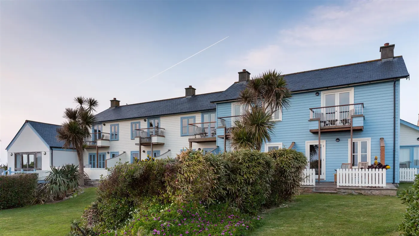 A front view of Hell Bay Hotel with charming blue and white coastal houses with balconies, surrounded by lush greenery and palm trees. A clear sky adds to the serene atmosphere.