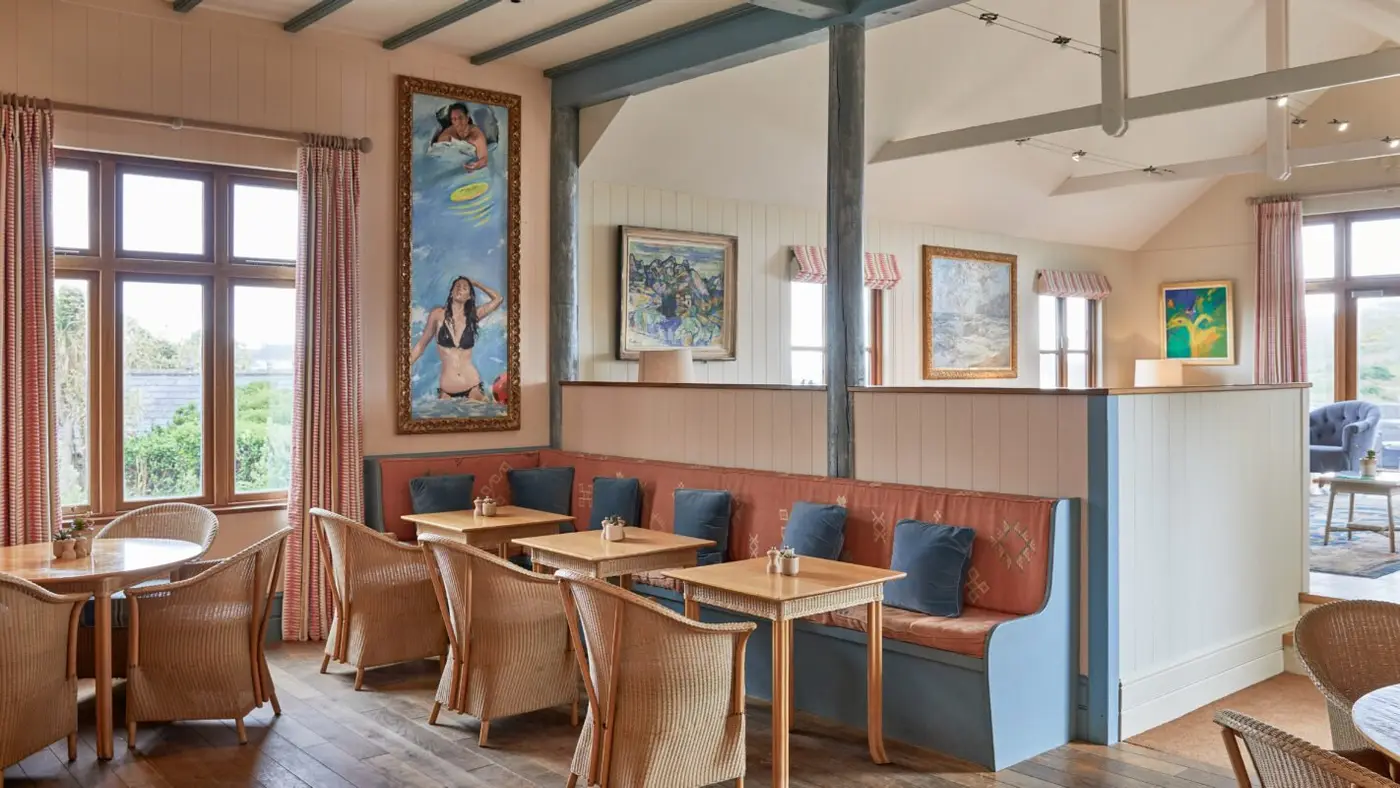 Cosy bar interior with wicker chairs and cushioned benches, featuring artwork on cream walls. Large windows with curtains let in natural light. Wooden flooring and beams add warmth to the space.