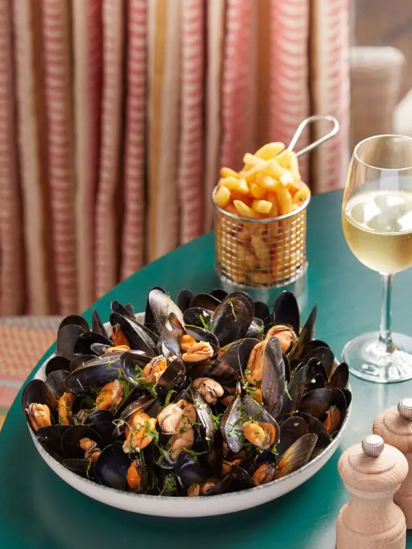 A bowl of steamed mussels garnished with herbs is on a green table. Behind it, a small basket of golden fries and a glass of white wine are placed. Striped curtains and salt/pepper shakers are visible in the background.