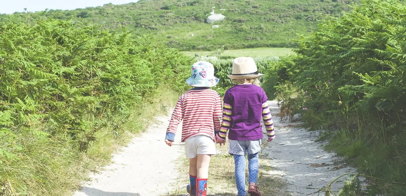 Two children wearing colourful clothing and hats walk hand in hand down a sandy track surrounded by green bushes. A hilly landscape is visible in the background under a clear sky.