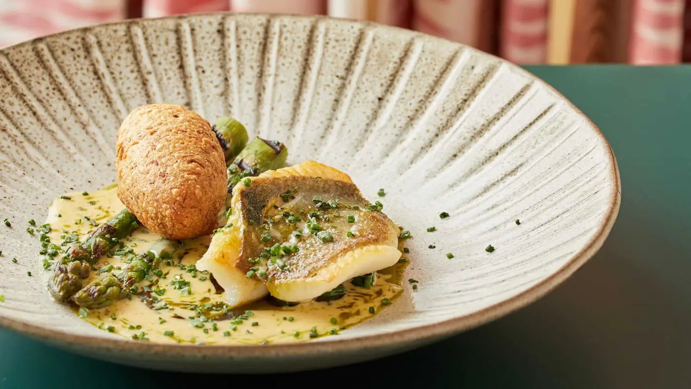 A dish featuring a fillet of fish with green herb sauce, garnished with finely chopped chives, next to grilled asparagus and a golden-brown croquette. Served on a textured beige plate with a green table background.