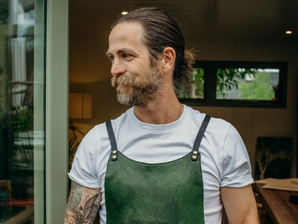 A man with a beard and mustache wears a white t-shirt and a green apron. He has a tattoo on his arm and is standing indoors, looking to the side and smiling.