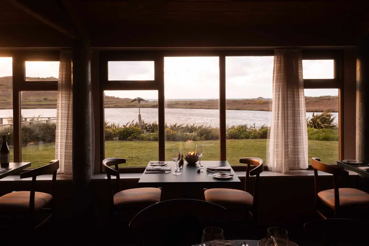 A cosy restaurant interior with wooden furniture and large windows offers a view of a serene lake and grassy landscape at sunset. Tables are set for two, with soft lighting creating a warm ambiance.