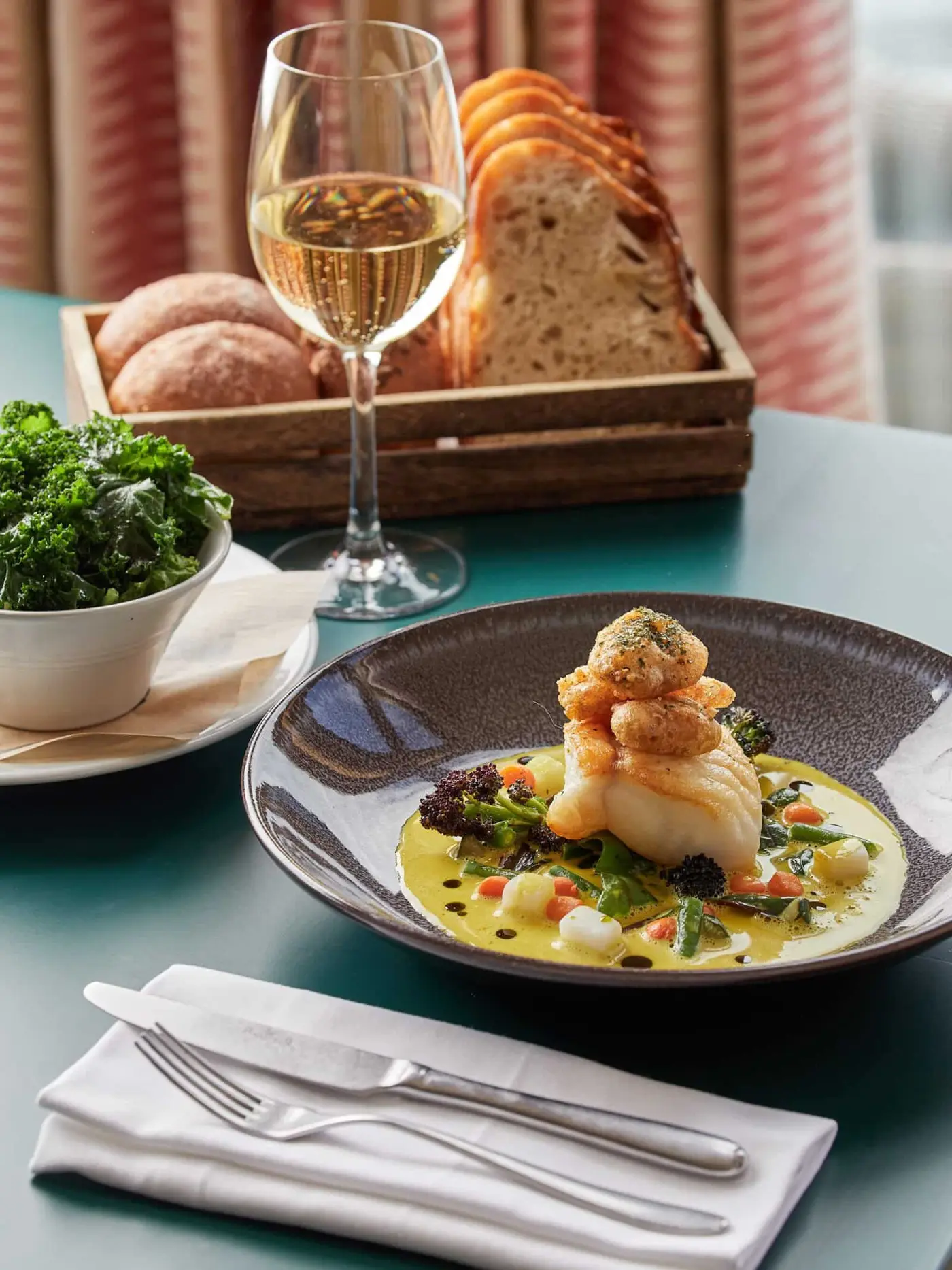 A gourmet dish featuring seared scallops atop white fish with a colorful vegetable medley and sauce. Accompanied by a bowl of leafy greens, a basket of bread, and a glass of white wine on a green table. Silverware and a napkin are nearby.
