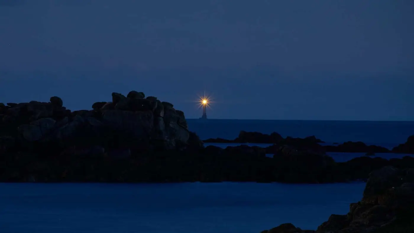 A distant lighthouse shines brightly against a dark blue night sky. Its light reflects off the calm ocean. Rocky formations are visible in the foreground, adding depth to the serene coastal scene.