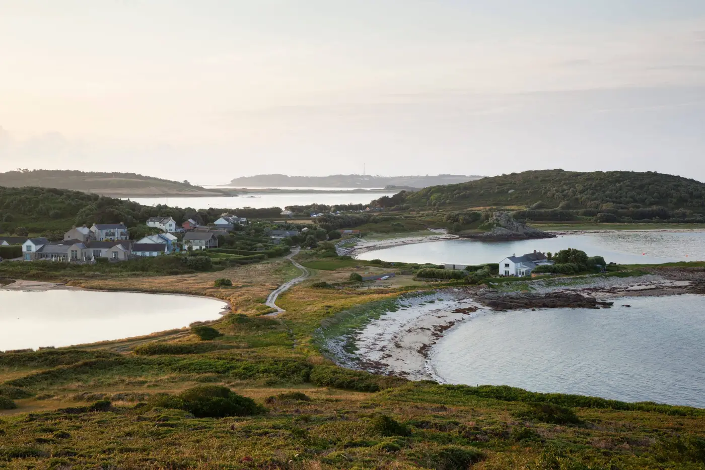 Scenic coastal landscape with grassy hills, small houses, and winding paths. Several inlets and a calm body of water are visible under a cloudy sky, creating a tranquil atmosphere.