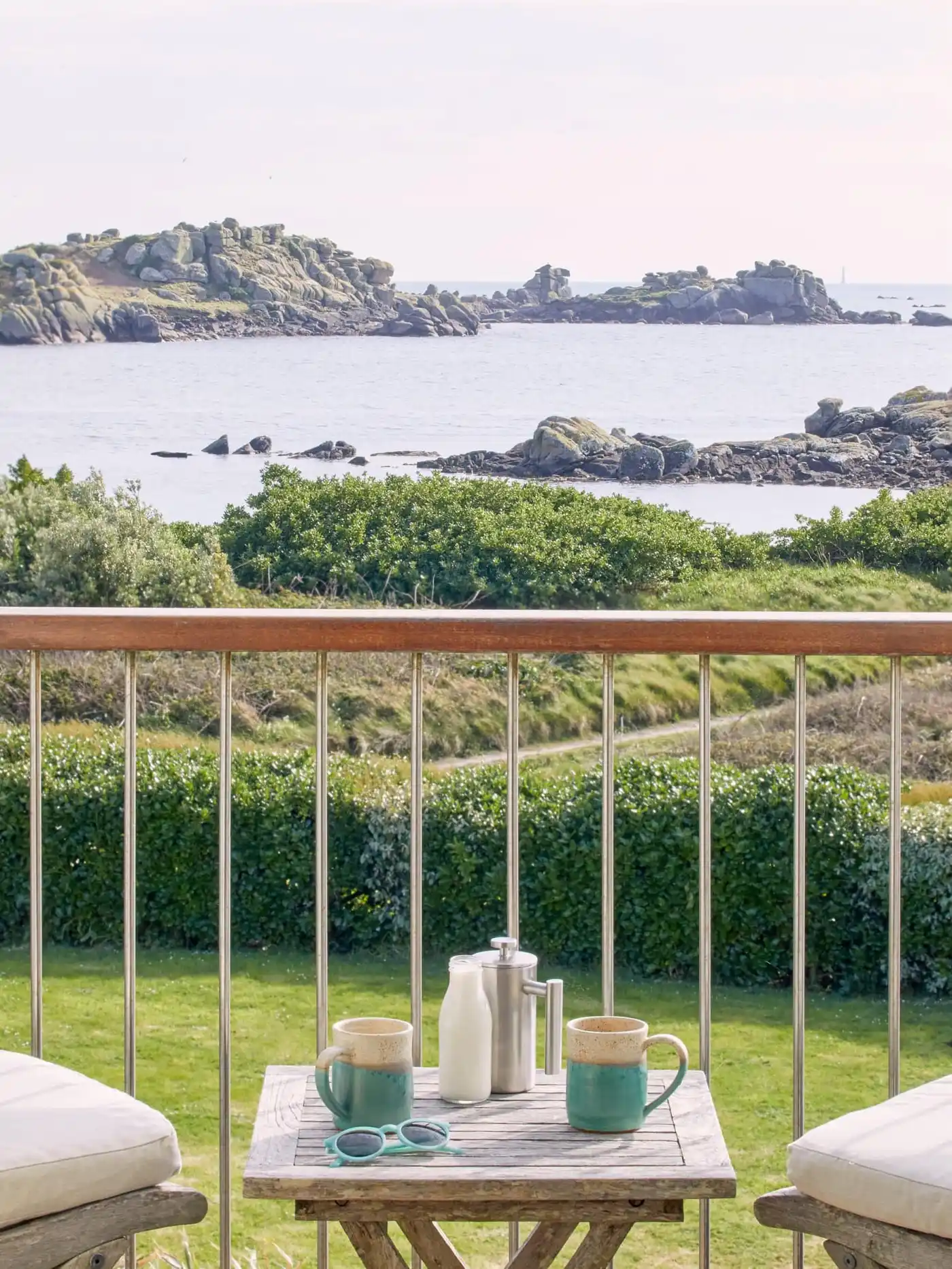 A scenic view of a rocky coastline with calm sea and green shrubs. In the foreground, a wooden table on a balcony holds two mugs, a milk jug, and a cafetiere. Two cushioned chairs are partially visible. Sunny and serene atmosphere.