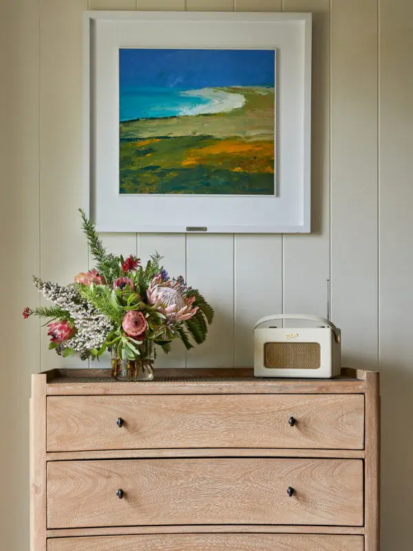A wooden dresser with three drawers holds a bouquet of flowers in a clear vase and a vintage-style radio. Above, a framed abstract landscape painting with blue and green hues hangs on the wall.