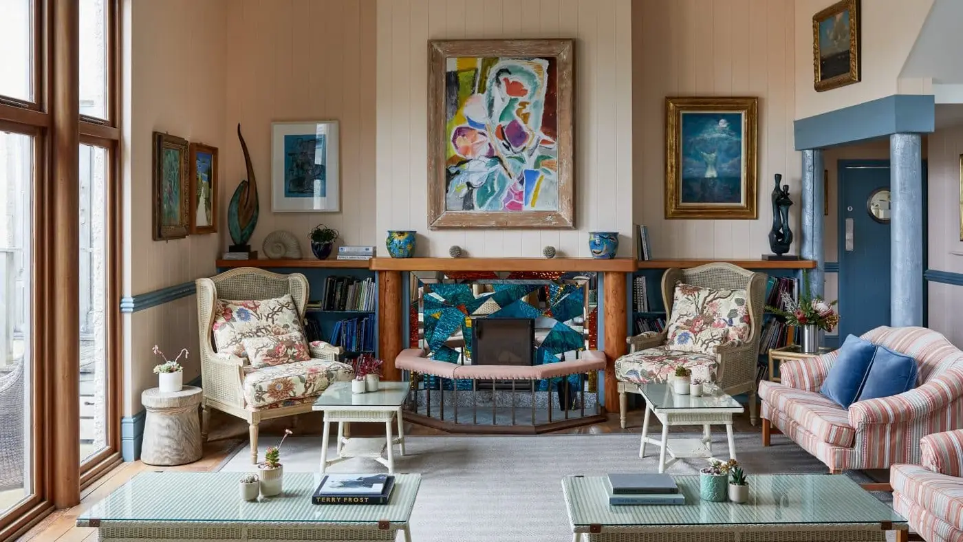 A cosy living room with eclectic décor, featuring wicker chairs with floral cushions, a striped sofa, and a colourful abstract painting above a mosaic fireplace. Large windows on the left bring in natural light, illuminating the space.