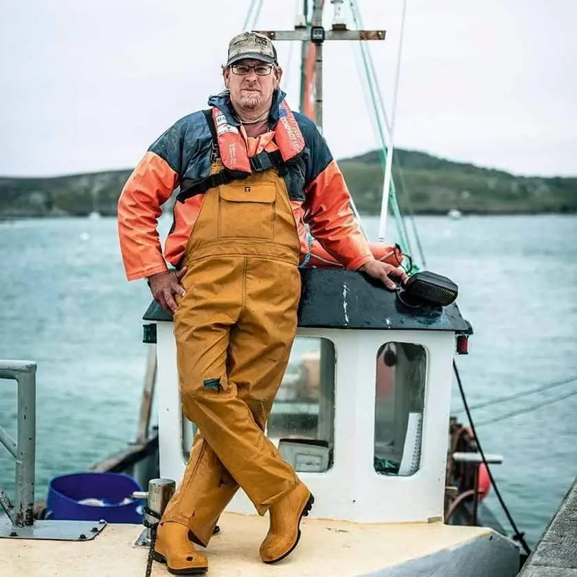 A fisherman - Mark Pender - wearing yellow overalls, an orange and blue jacket, and a cap leans against the cabin of a small fishing boat. The background shows a calm body of water and distant hills under an overcast sky.