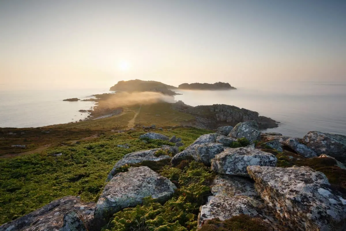Sunset view of a rocky coastline over the north end of Bryher, with Shipman Head in the distance. The sun is low, casting warm light over the water and rocks. Patches of green vegetation are visible in the foreground, with a misty atmosphere.