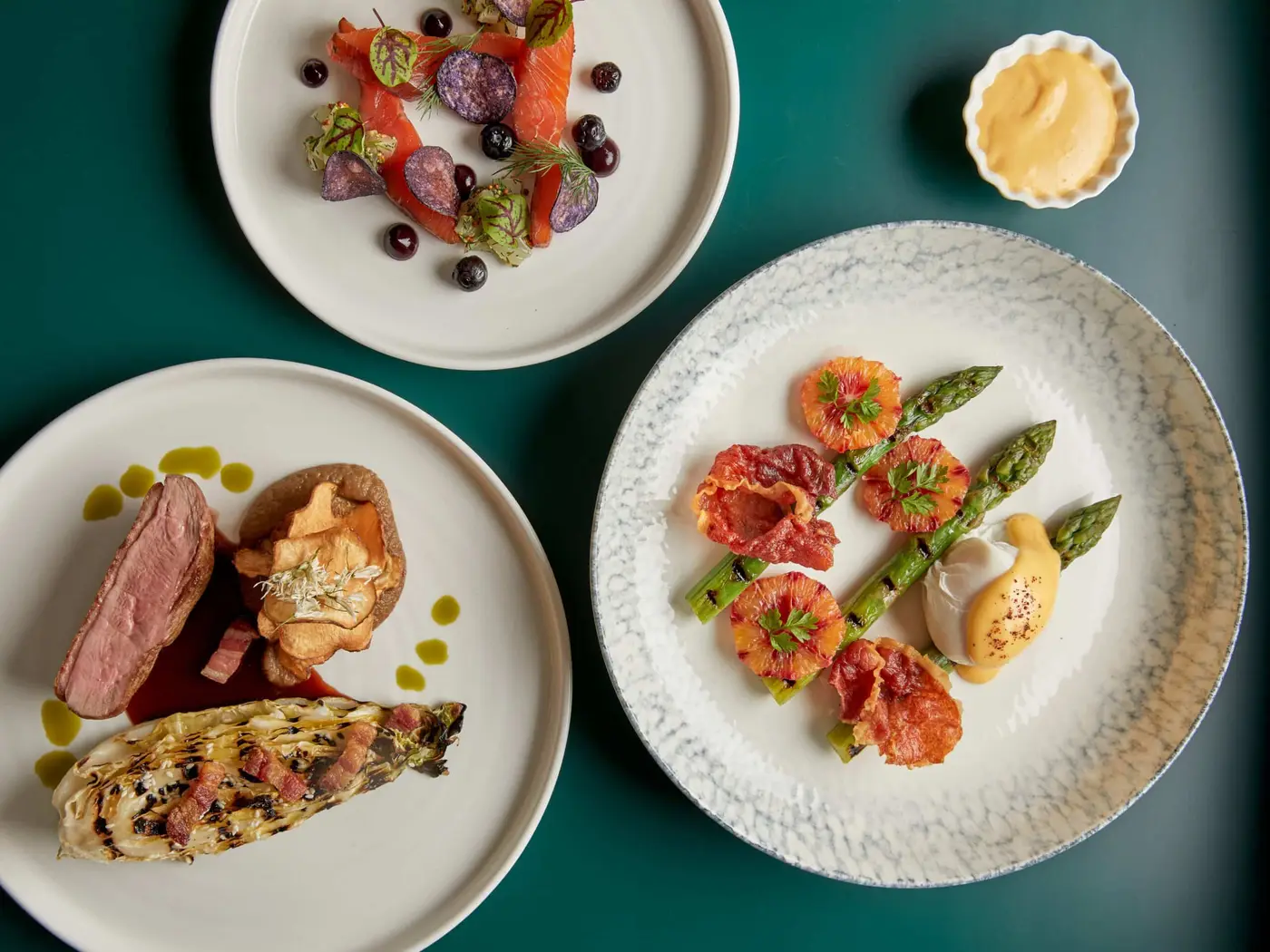 Aerial view of three elegantly plated dishes on a green table. Top left: vibrant salad with berries and herbs. Bottom left: steak with Yorkshire pudding and grilled lettuce. Right: asparagus with crispy vegetables and a dollop of creamy sauce.